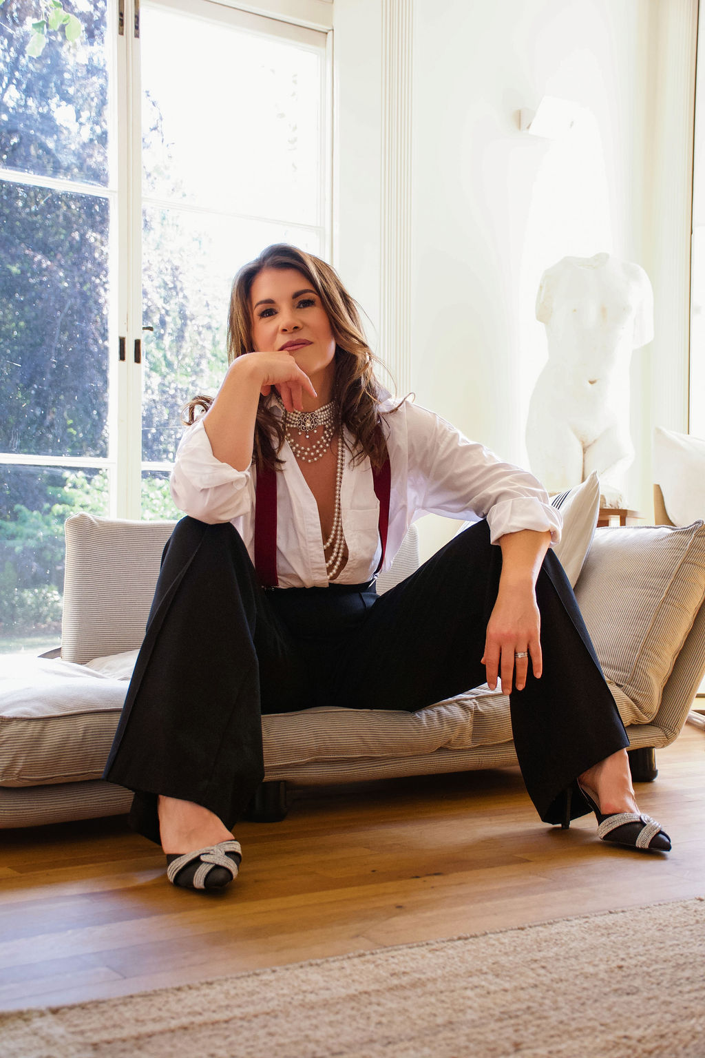 Samantha Charlwood seated on a sofa in a white shirt, pearls and black trousers, looking directly at the camera.